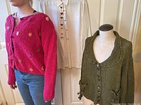 Model wearing pink hand knit floral embroidered sweater and showing olive green hand knit sweater on mannequin with Todd Oldham jeweled dress shirt behind.