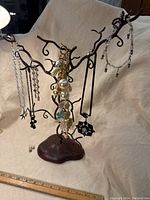 Brown metal display tree with multiple necklaces hung on its branches; visible are necklaces with large faux pearls, black web pendant, and clear bead chains.