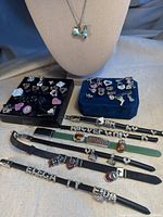 Photo showing assortment of vintage charm bracelets on metal and leather bands with various charms including hearts, letters, keys, and novelty shapes, arranged on fabric surface with black and blue display trays.