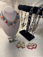 Lot of colorful whimsical vintage jewelry including necklace displayed on stand, multi-strand necklaces on jewelry bar, two pairs of earrings and brooch on fabric surface.