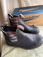Side view of pair of black leather Blundstone boots with multicolored striped elastic side panels, placed on top of the Blundstone box.