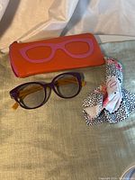 Photo showing the purple and yellow Caroline Abram prescription glasses alongside an orange zippered case with pink glasses silhouette, and a polka-dotted cloth for cleaning.