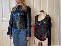 Model wearing the black leather jacket, standing next to a mannequin with the patterned top. Both items displayed indoors against white doors.