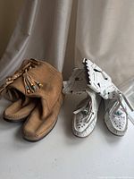 Two pairs of moccasin boots, one in light brown suede with bead-tipped tassels, the other in white leather with cut-outs and stud details.