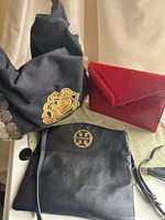 Overview of three vintage purses together on cloth background under natural light. Includes black Tory Burch leather purse, black fabric purse with gold details, and red hardcase clutch.