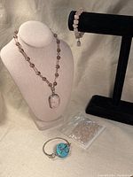 Full view of the three main jewelry pieces including rose stone pendant necklace, rose stone bead bracelet on a display, turquoise stone wire bracelet, and the small bag of loose rose stone beads.