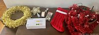 Top view of all Christmas items laid on table: gold garland wreath, indoor lights, star vintage style tree topper, stockin,g red garland, and woven basket