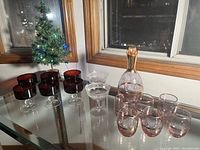 Photo showing the purple Spirit decanter with gold top, six purple matching glasses, two clear etched coupes, and six brown Luminarc dessert/coupes with clear stems on a glass table near a window with a small lit Christmas tree.