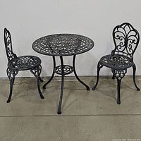 Full view of the complete 3-piece wrought iron bistro set showing one table and two chairs on concrete floor.