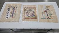 Photo showing all three Egyptian papyrus paintings laid out on a table, each wrapped in plastic, with visible Egyptian figures and hieroglyphs.