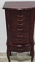 Front view of the dark wood Queen Anne style jewelry armoire showing eight drawers with brass handles.
