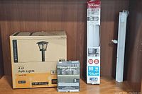 Wide shot of four lighting items: boxed solar LED path lights, boxed 18-inch LED under cabinet light, LED tape lights coil, and unboxed light fixture