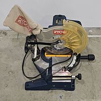 Front angle view of Ryobi compound miter saw showing dust collection bag, yellow blade guard, blue body, and cord, placed on concrete floor.