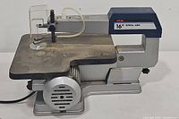 Front and side view showing the scroll saw's metal work table, motor housing, power cord, and Ryobi branding on the blue upper arm.