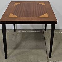 Front view of vintage wooden card table showing marquetry patterned top with corner inlays and four dark-stained legs.
