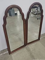 Pair of vertical wall mirrors with wooden frames and scalloped tops, shown together leaning against a wall in a warehouse space.