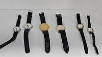 Six wristwatches laid out in a row showing variety of dial designs, band types, some with black leather bands and some with black elastic bands including two Mickey Mouse designs.