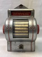 Front view of the jukebox wallbox showing silver metal body, red knobs on sides, display window with music selections, and small slot at bottom.