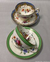 Two teacup and saucer sets shown together on a flat surface; one with green band and floral design, the other with blue and gold floral pattern.