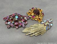Lot of three vintage rhinestone brooches arranged on gray surface; shows color, cut, and metal details.