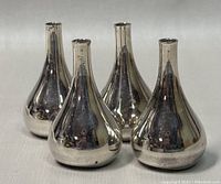 Four polished metal teardrop candleholders with narrow necks and reflective surfaces arranged on a white cloth.