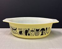 Side angled view of Pyrex dish showing yellow exterior with black Mod Kitchen vegetable and fruit graphics, white interior, and handle.