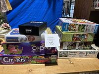Photo showing stacked board games 'World's Smallest Party Game', 'Cadoo', 'Cir-Kis', 'Blockbuster', and three 1000-piece puzzles including 'Summer Cottage'.