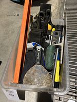 Top down view of clear plastic bin holding multiple used hand tools such as putty knife, holster with scissors, digital thermometer, and flashlight