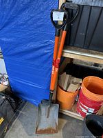Two metal shovels leaning against shelving, both with orange shafts and black D-grip handles. Both show rust on the flat metal blades.