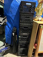 Stack of six black plastic milk crates with lattice design, showing rectangular hand grips and some white text and numbers printed on sides, dusty condition.