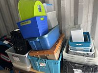 Plastic bins and totes stacked including blue, transparent, black, gray, beige bins and green lid