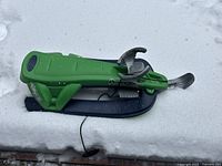 Side view of green plastic snow racer sled on snow showing steering handle and foot brake.