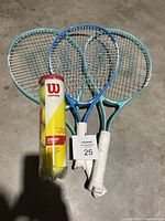 Three Wilson tennis rackets with one can of Wilson tennis balls shown on concrete floor. Two rackets are teal color, one is blue.
