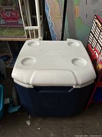Exterior front and top view of blue rectangular Coleman cooler with white lid featuring four cup holders and large black wheels on the side.