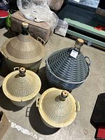 Overview of four wine making carboys in wicker and plastic protective covers with cork stoppers on a concrete floor.