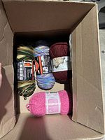 Top-down view of four balls of yarn inside a cardboard box. Showing pink Bernat Blanket yarn, multi-colored, green-yellow-brown variegated yarn, and a dark red yarn ball.