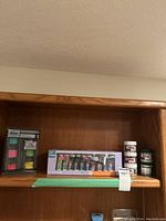 Wooden shelf displaying various acrylic art supplies including paint tubes and jars of texture pastes and glazes.