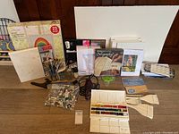 Wide view of mostly all items together on the floor showing watercolour trays, brushes, calligraphy set, paint tubes, paper pads, small easel, and more.
