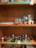Photo of two shelves in a wooden unit showing a variety of art supplies including acrylic paint tubes, pigment jars, paint markers, brushes, paint mediums, and glitter jars.