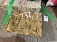 Seven glass blown icicle ornaments and one small decorative glass tree arranged on a gold patterned cloth with tassels, displayed on wooden floor near brick.