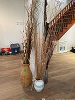 Full view of three vases standing on floor filled with tall decorative dried grasses and sticks in a home setting near wooden stairs.