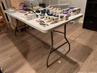 White 6 ft folding table set up on wooden floor with various items on top (items not included in sale). Table legs are foldable metal.