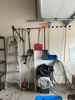 Photo showing a silver aluminum step ladder next to a wall rack with hanging garden tools including rakes, shovels, brooms, and plastic shovels, along with black and white tarps folded underneath and a coiled metal hose on the floor.