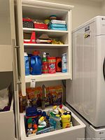 Opened kitchen cabinet and lower drawer with various household cleaning products including laundry detergent bottles, detergent sachets, cleaning sprays, and other supplies.
