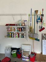Wall-mounted white shelves holding multiple bottles of cleaning products including sprays, liquid detergent, and surface cleaners. Buckets of various colors at the floor below. Cleaning tools hanging on hook rack beside the shelves.
