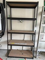 Front view of five-shelf industrial metal shelving unit showing particleboard shelves with visible dirt and wear spots, black metal perforated frame.