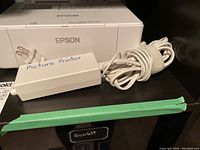 Epson PictureMate PM-400 printer with connected power adapter and some photo paper in the background.