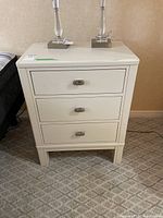 Front view of white nightstand with three drawers and two lamps on top