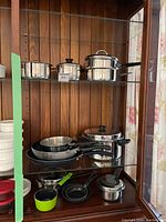 View of assorted pots and pans arranged on glass shelving, including stainless steel pots with lids and various frying pans.