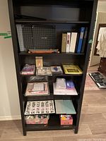 Photo of black shelving unit with scrapbooking supplies including three paper cutters, binders of various colors, alphabet stickers, crafting embellishments, and tools.